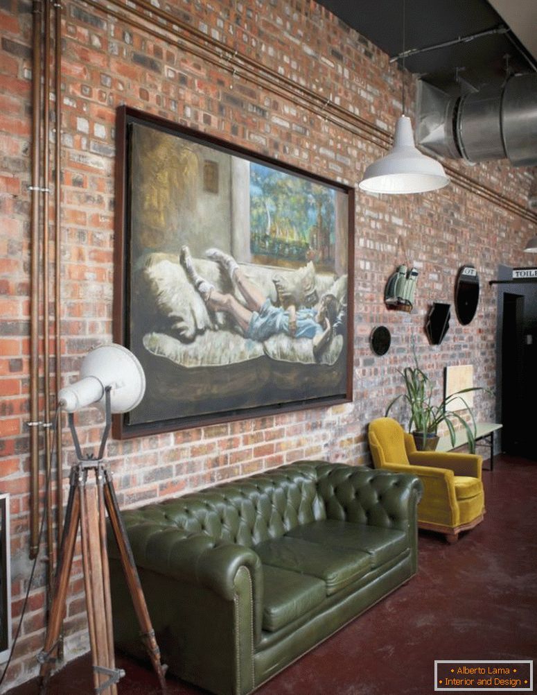Large oil painting on brick wall of industrial-style loft apartment with retro sofa and armchair